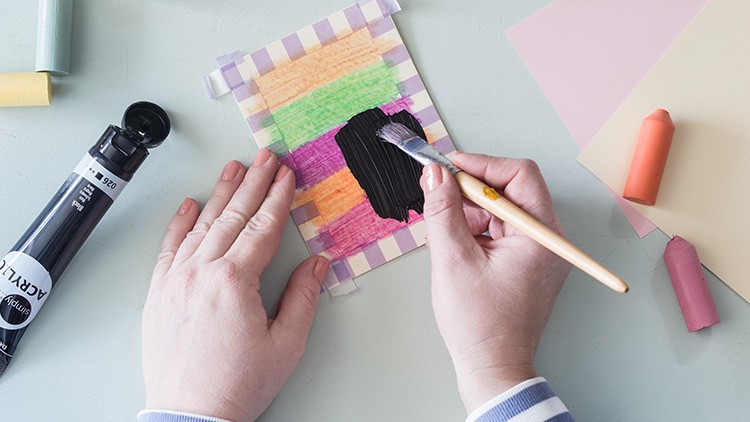 Eine Hand malt mit einem Pinsel schwarze Farbe auf ein buntes Papier, daneben liegen Pastellkreiden und eine schwarze Acrylfarbe.