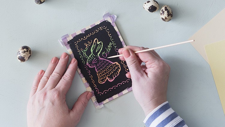 Eine Hand gestaltet mit einem Holzstab ein buntes Kratzbild, daneben liegen Wachteleier auf einem hellen Hintergrund.