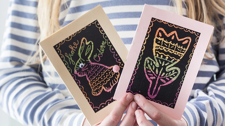 Zwei bunt gestaltete Karten mit Osterhasen- und Blumenmotiv werden von einer Person in gestreiftem Oberteil gehalten.