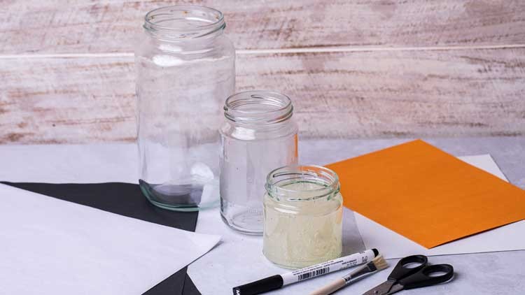 Drei leere Glasgefäße stehen auf Bastelmaterialien wie farbigem Papier, Schere, Pinsel, Kleber und einem schwarzen Filzstift.