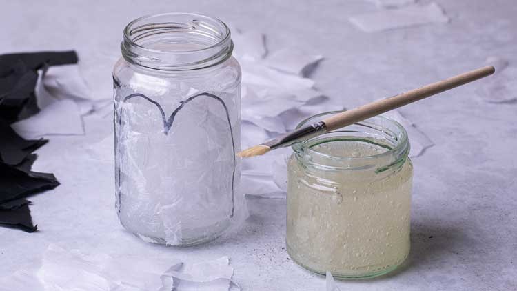 Ein leerer Glasbehälter mit weißem Papier beklebt, daneben ein kleiner Glasbehälter mit Kleber und ein Pinsel auf grauem Untergrund.