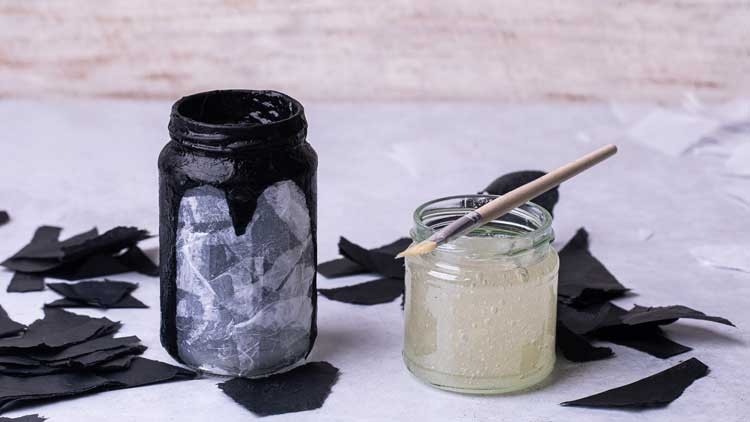 Ein schwarzes Glas mit halbtransparenten Papierstücken steht neben einem Glas mit Kleber und Pinsel, umgeben von Papierschnipseln.