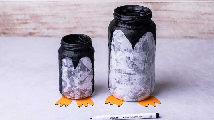 Zwei bemalte Gläser in Pinguinform mit schwarzen, weißen und orangefarbenen Details neben einem weißen STAEDTLER Whiteboard-Marker.