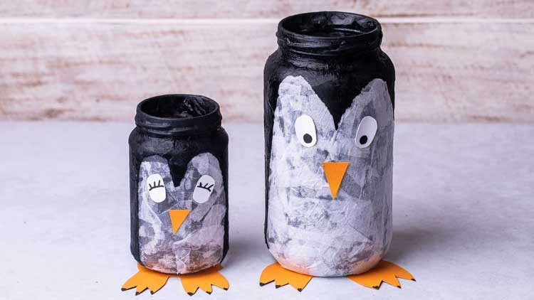 Zwei bemalte Gläser in Form von Pinguinen mit Papieraugen, orangefarbenen Schnäbeln und Füßen stehen auf hellem Untergrund.