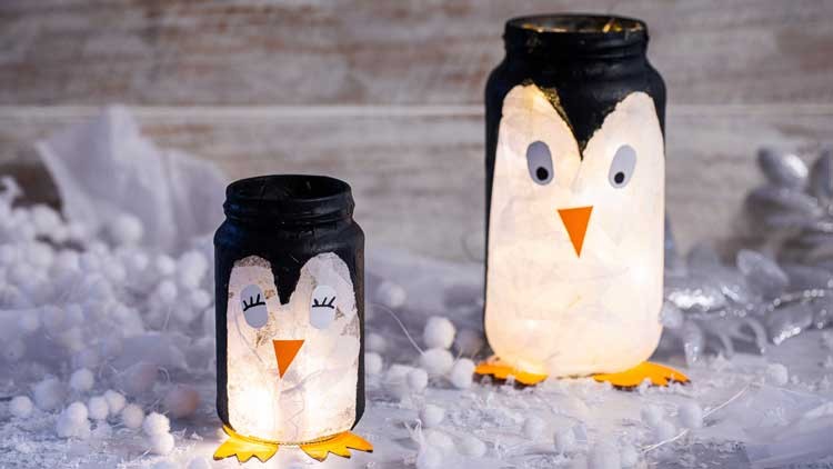 Zwei dekorierte Einmachgläser in Pinguinform mit leuchtender Füllung auf Winterdekoration mit weißen Kugeln und Stoff.