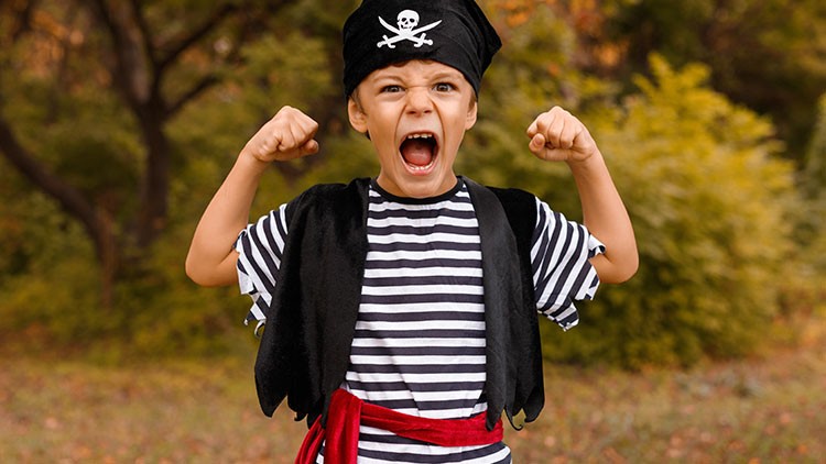Person in schwarz-weiß gestreiftem Oberteil, rotem Gürtel und Piratenhut mit Totenkopf steht draußen und zeigt geballte Fäuste.