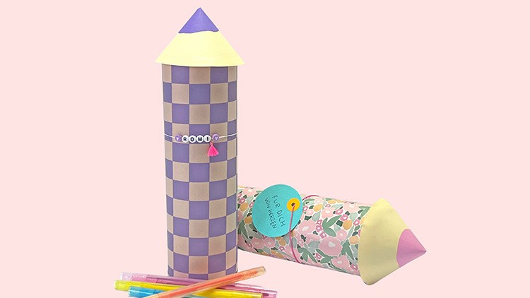 Zwei bunt verzierte Raketen aus Papier mit gemusterten Hülsen und Kegeln, daneben liegen bunte Filzstifte auf hellem Hintergrund.