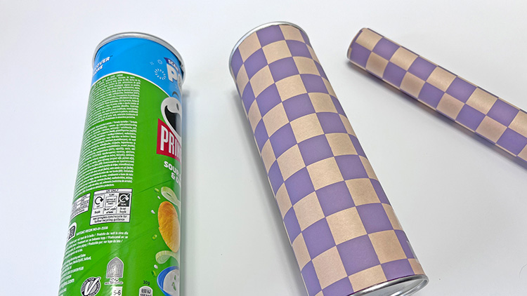 Eine Chipsdose mit grünem Design und zwei Dosen mit lila-beigem Schachbrettmuster liegen auf einem weißen Untergrund.