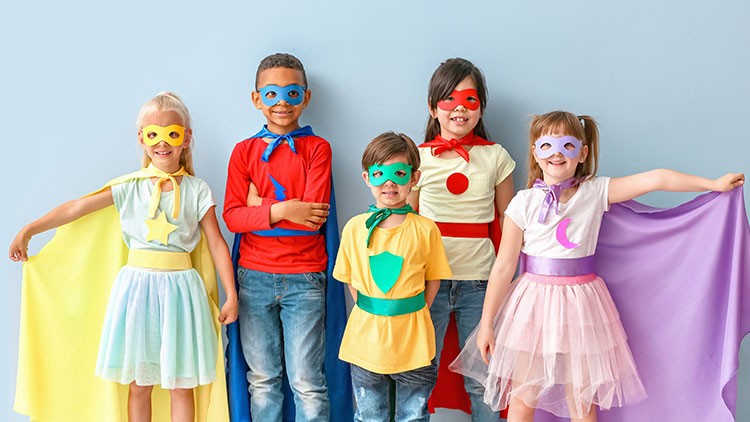 Fünf Kinder in bunten Superheldenkostümen mit Umhängen vor einer hellblauen Wand.