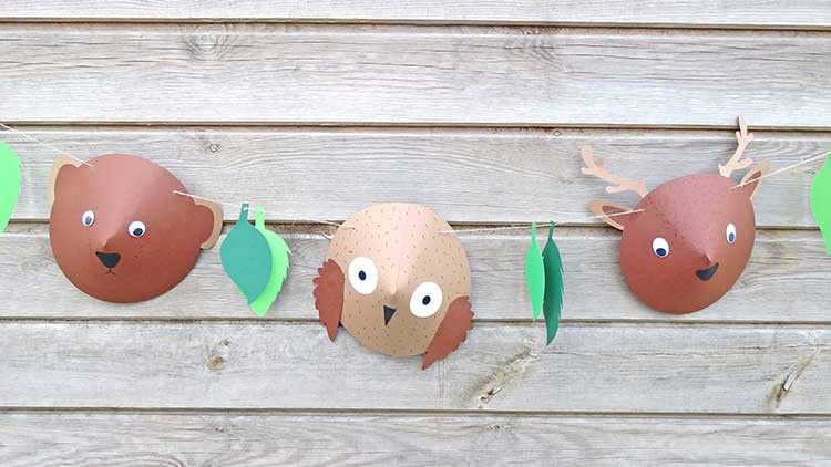 Girlande aus Pappfiguren eines Bärs, einer Eule und eines Hirsches mit grünen Papierblättern an Holzwand befestigt.