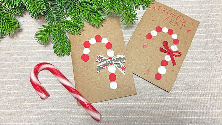 Zwei braune Karten mit roten und weißen Zuckerstangenmotiven, Zweig und Zuckerstange aus Kunststoff auf gestreifter Tischdecke.