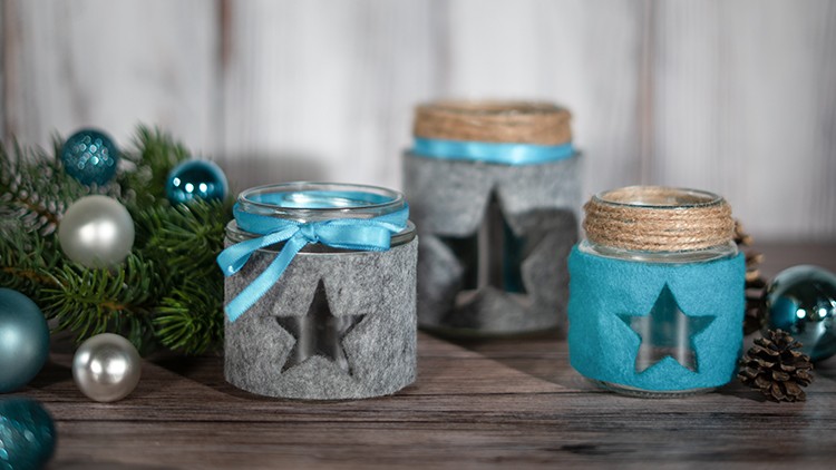Drei Gläser mit Sternmustern aus Filz, dekoriert mit Schleifen und Naturfaser, umgeben von Tannenzweigen und Kugeln.
