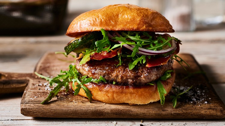 Burger mit Rindfleisch-Patty, Rucola, roten Zwiebelringen, gebratenem Speck und Gemüse auf einem Holzbrett.