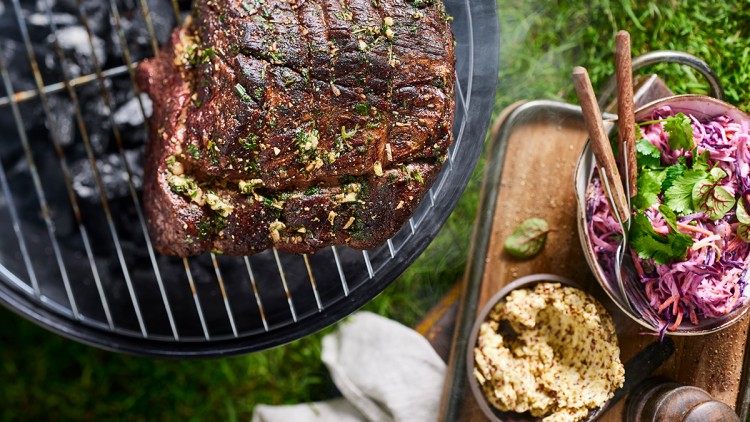 Ein gegrilltes Stück Fleisch mit Kräutern auf einem runden Grill neben Krautsalat, Senf und einem Holzbrett auf grünem Rasen.