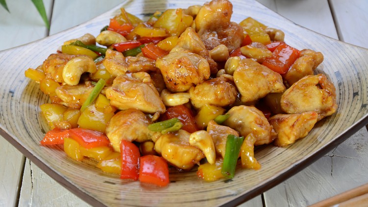 Gebratene Hähnchenstücke mit Paprika, Lauchzwiebeln, Cashewkernen und Sauce auf einem beigen Teller.