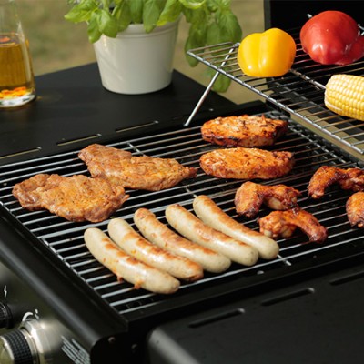 Auf einem Grill liegen Würstchen, Steaks, Hähnchenflügel und Maiskolben, daneben gelbe und rote Paprika sowie frisches Basilikum.