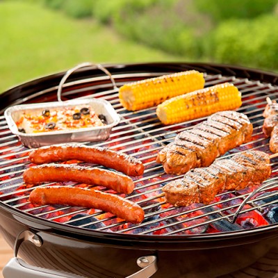 Verschiedene Grillgerichte wie Würstchen, Fleisch, Maiskolben und eine Gemüseauflauf-Schale liegen auf einem runden Holzkohlegrill.