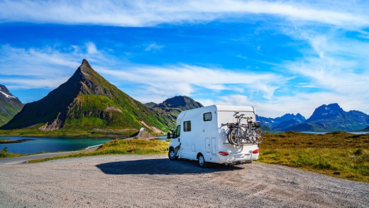 Ein weißes Wohnmobil mit zwei Fahrrädern am Heck steht auf einem Parkplatz vor einer Berglandschaft mit See und blauem Himmel.