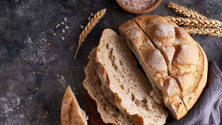 Ein angeschnittenes rundes Brot mit knuspriger Kruste liegt auf einer dunklen Fläche mit Weizenähren und Salz.