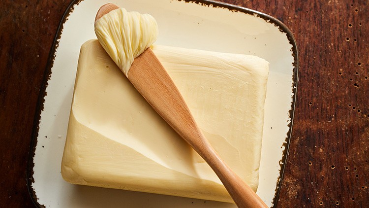 Ein Stück Butter liegt auf einer weißen Keramikplatte, ein Holzmesser mit Butter darauf liegt schräg auf der Butter.