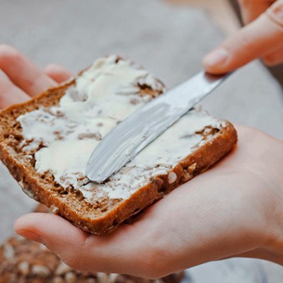 Eine Hand streicht mit einem Messer Butter auf ein Scheibe Vollkornbrot, im Hintergrund ein Glas mit Joghurt und Beeren.