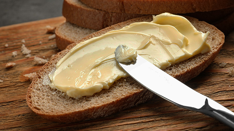 Ein Messer streicht gelbe Butter auf eine Scheibe braunes Brot, das auf einem rustikalen Holztisch liegt.