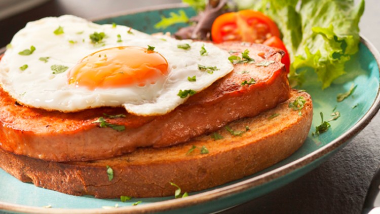 Ein gebratenes Spiegelei liegt auf gebratenem Fleisch und einer Scheibe Brot, daneben Salat und Tomate auf einem grünen Teller.