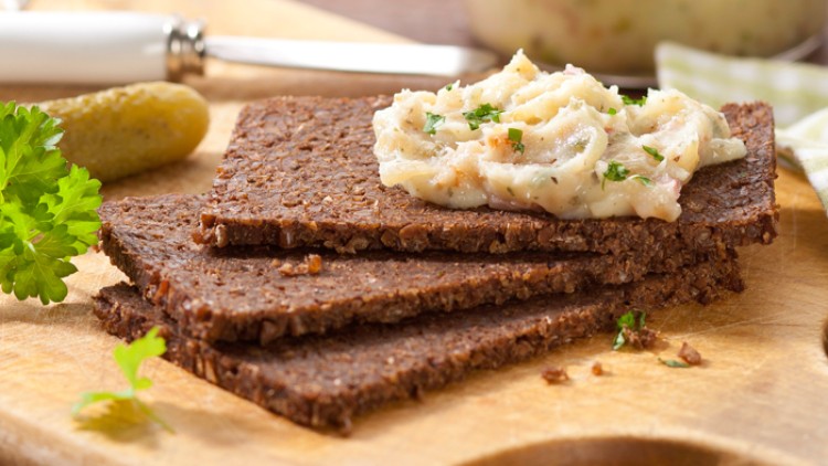 Drei Scheiben dunkles Vollkornbrot mit cremigem Aufstrich, dekoriert mit Kräutern, auf Holzbrett neben Gurke und Petersilie.
