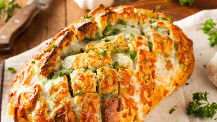 Grob geritztes, überbackenes Brot mit geschmolzenem Käse, Kräutern und Knoblauch auf einem Holzbrett und einem Tuch.