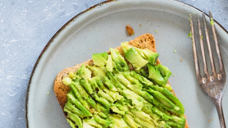 Getoastete Brotscheibe mit zerdrückter Avocado auf einem hellgrauen Teller neben einer silberfarbenen Gabel.