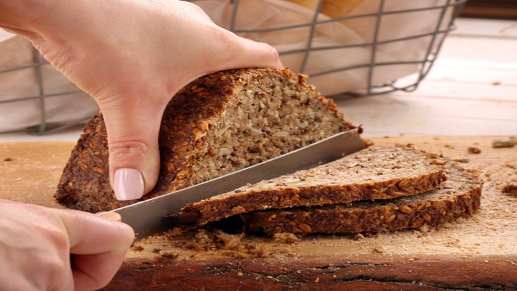 Eine Hand schneidet mit einem Messer zwei Scheiben körniges Vollkornbrot von einem Laib auf einem Holzbrett ab.