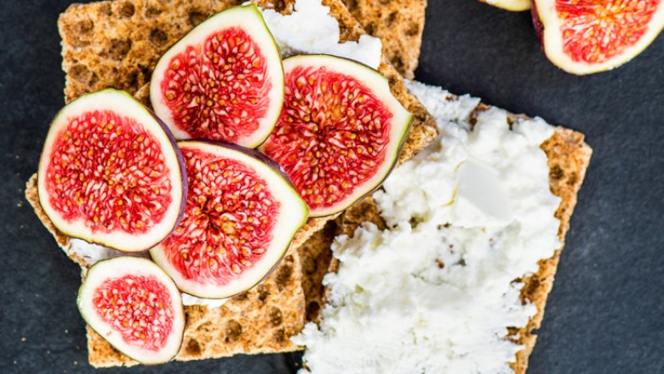 Knuspriges Knäckebrot mit weißem Frischkäse und frischen, aufgeschnittenen Feigen auf dunklem Untergrund.