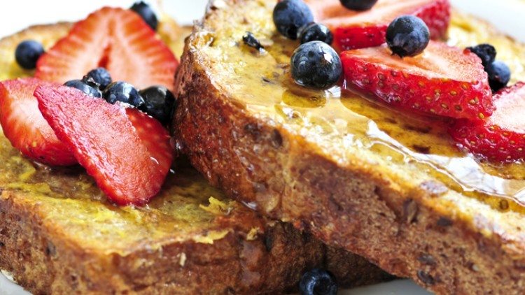Zwei Scheiben French Toast belegt mit frischen Erdbeeren, Blaubeeren und überzogen mit flüssigem Sirup auf einem weißen Teller.