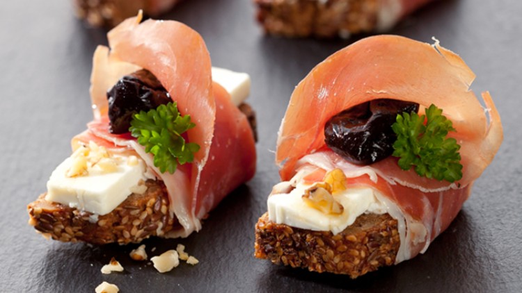 Häppchen aus Vollkornbrot mit Frischkäse, Schinken, Pflaume, Walnuss und Petersilie auf schwarzer Platte angerichtet.