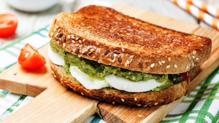 Getoastetes Vollkorn-Sandwich mit Mozzarella, grünem Pesto und Tomaten auf einem Holzbrett, daneben halbierte Kirschtomaten.