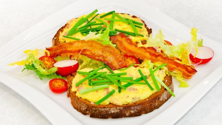 Zwei Scheiben Brot mit Rührei, Schnittlauch und knusprigem Speck, garniert mit Salat, Tomaten und Radieschen auf einem weißen Teller.