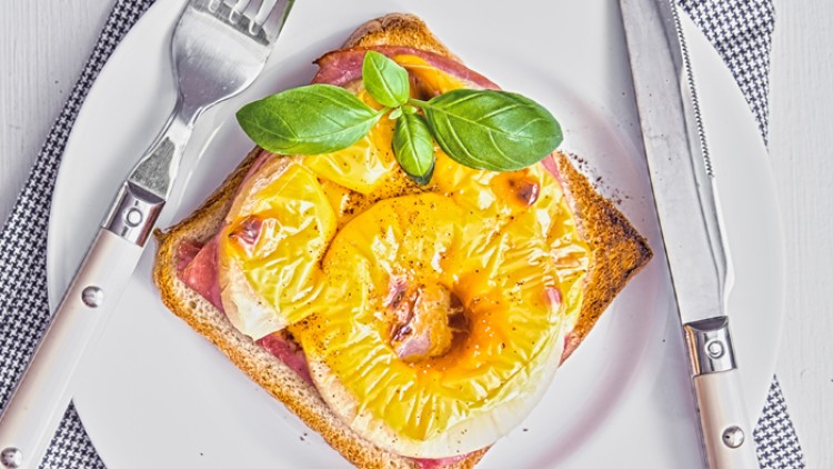Toast mit Schinken, geschmolzenem Käse, Ananasring und Basilikumblättern auf einem weißen Teller mit Messer und Gabel.