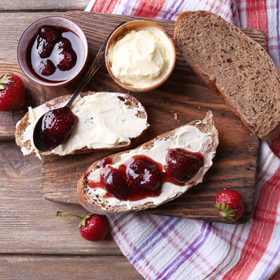 Ein Holztisch mit zwei bestrichenen Brotscheiben, roter Erdbeermarmelade, Butter, frischen Erdbeeren und einem karierten Tuch.