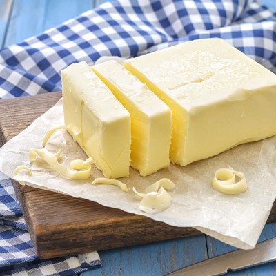 Ein Block Butter mit drei angeschnittenen Scheiben liegt auf Backpapier auf einem Holzbrett, daneben ein blau-weiß kariertes Tuch.