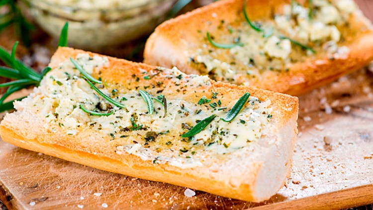 Zwei Halbstücke knuspriges Baguette mit Kräuterbutter und frischem Rosmarin auf einem hölzernen Schneidebrett.