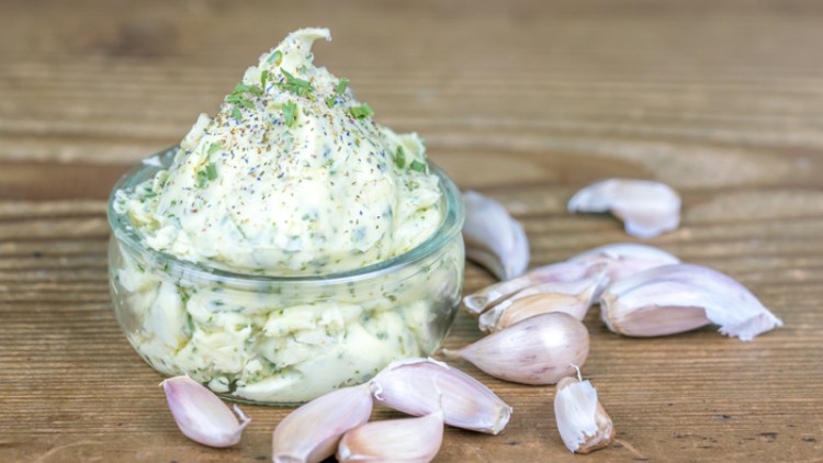 Kleines Glas mit Kräuterbutter, dekoriert mit Kräutern, umgeben von mehreren ungepulten Knoblauchzehen auf Holztisch.