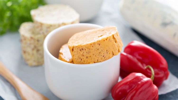 Rote Paprika, Kräuterbutterscheiben in einer weißen Schale, Holzlöffel und Kräuter auf hellem Untergrund.