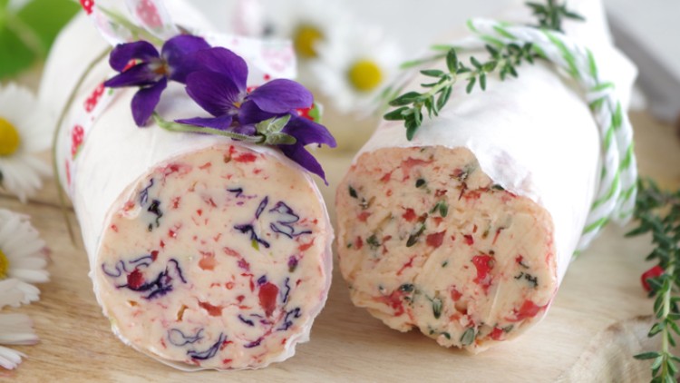 Zwei Kräuterbutterrollen mit Blüten und Kräutern dekoriert auf einem Holzbrett, umgeben von Gänseblümchen.