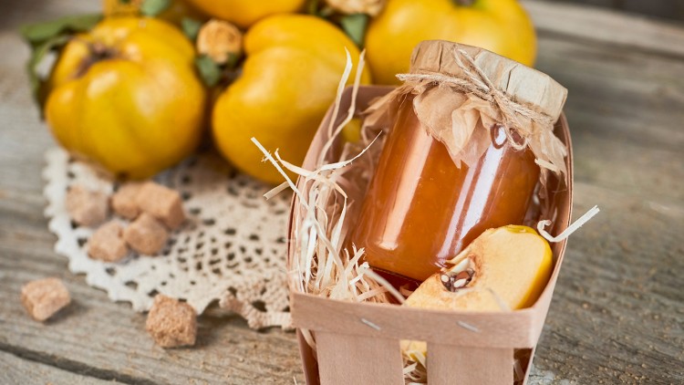 Ein Glas mit orangefarbener Marmelade steht in einer Holzschale mit Stroh, daneben Quitten und braune Würfelzucker auf einem Holztisch.