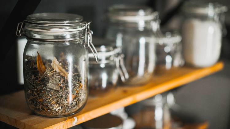 Mehrere verschlossene Glasbehälter mit Trockenkräutern und Pulver stehen auf einem Holzregal in einem Innenraum.