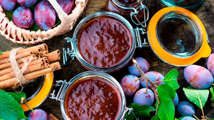 Offene Marmeladengläser mit Pflaumenmus, frische Pflaumen, Zimtstangen und grünen Blättern auf einem rustikalen Holzuntergrund.