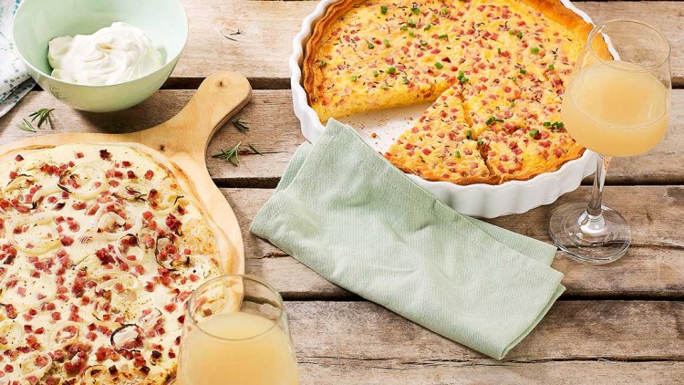 Flammkuchen und angeschnittene Quiche mit Speckwürfeln, zwei Gläser hellem Getränk, Schüssel mit Creme auf Holztisch.