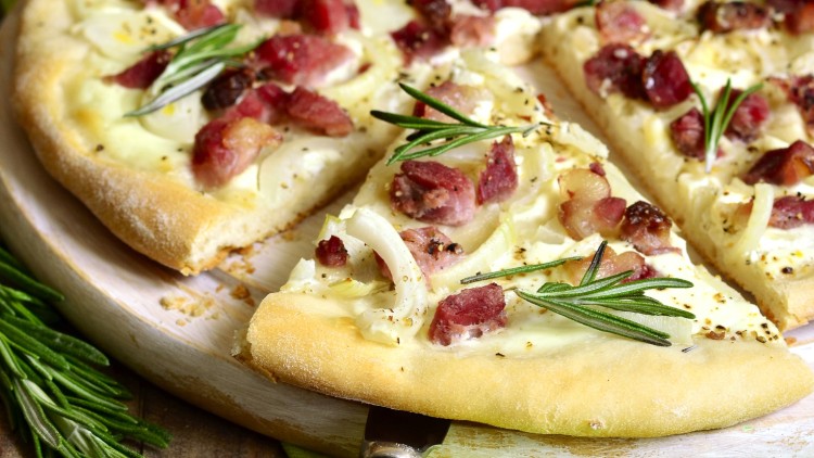 Flammkuchen mit Speckwürfeln, Zwiebeln, Rosmarinzweigen und heller Creme auf einem runden Holzbrett.