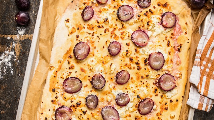 Flammkuchen mit halbierten roten Trauben und zerbröckeltem Käse auf Backpapier, daneben Holzuntergrund und Stoffserviette.
