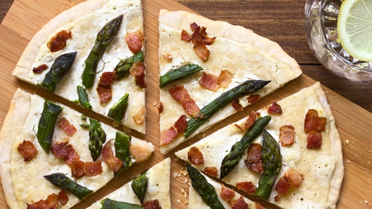 Pizzastücke mit weißer Sauce, grünem Spargel und gebratenen Speckwürfeln auf Holzbrett, daneben Glas Wasser mit Zitronenscheibe.